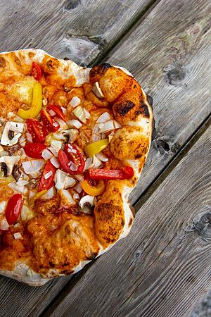 Eine ausgebackene Pizza, belegt mit frischen Champignons, Paprika, Zwiebeln und Tomatensauce, liegt auf einem rustikalen Holztisch.