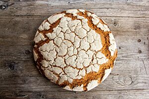 Hofbrot. Ein großer, runder Laib mit bemehlter, rustikaler Kruste liegt auf einem grauen Holztisch.