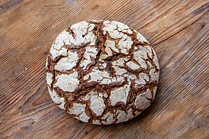 Helles Roggenbrot. Ein runder, dunkel ausgebackener Laib mit bemehlter, rustikaler Kruste liegt auf einem braunen Holztisch.