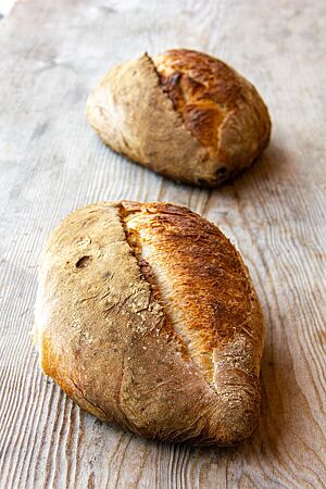 Französisches Landbrot. Zwei längliche, spitz zulaufende Laibe mit mittelbraun ausgebackener, bemehlter Kruste liegen auf einem Holztisch.