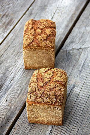 Pumperbrot. Zwei im Kasten gebackene Pumperbrote mit Schrot und vielen Rissen auf der Kruste liegen auf einem rustikalen Holztisch.