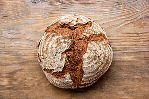 Dinkelvollkornsauerteigbrot mit Buchweizen. Ein runder Laib mit rustikal aufgerissener, bemehlter Kruste liegt auf einem hellen Holztisch.