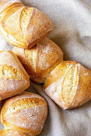 Frühstücksbrötchen. Die goldgelb ausgebackenen, rechteckigen Frühstücksbrötchen mit diagonalem Einschnitt und bemehlter Kruste liegen auf einem grauen Leintuch.