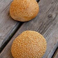 Zwei goldgelb ausgebackene Burger Buns mit Sesam auf der glatten Oberfläche liegen auf einem rustikalen Holztisch.