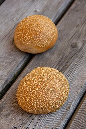 Zwei goldgelb ausgebackene Burger Buns mit Sesam auf der glatten Oberfläche liegen auf einem rustikalen Holztisch.