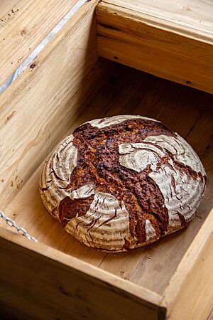 Roggenbrot mit Malzstück. Das kräftig ausgebackene, rustikal aufgerissene Roggenbrot mit bemehlter Kruste liegt in einer Holzkiste.