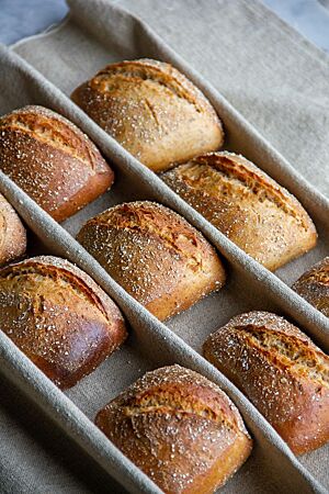 Dreikornkrustis. Eckige, rustikal aufgerissene Brötchen mit Schrot auf der Kruste liegen in Reihen auf einem hellen Leinentuch.