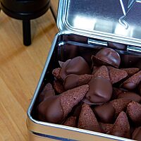 Die braunen, am breiten Ende mit Schokolade überzogenen Neidarfer Raacherkarzle liegen in einer silbernen Dose aus Metall.