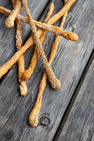 Grissini. Lange, dünne Grissini-Stangen mit hellbrauner Kruste und aufgestreutem Sesam liegen auf einem grauen Holztisch.