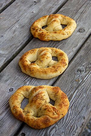Dinkel-Pfeffer-Brezen. Drei dicke hellbraune Brezeln mit gemahlenem schwarzem Pfeffer auf der Kruste liegen auf einem grauen Holztisch.