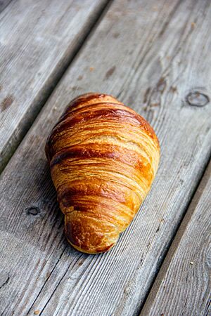 Ein goldbraun ausgebackenes Croissant mit leicht glänzender Oberfläche liegt auf einer rustikalen Holzbank.