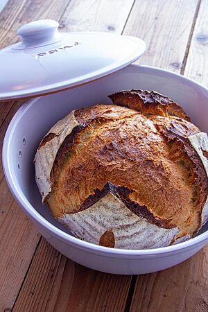 Römertopfbrot. Das kräftig ausgebackene, weit aufgerissene Römertopfbrot mit bemehlter Kruste und großem Volumen liegt in einem weißen Brottopf mit Deckel.