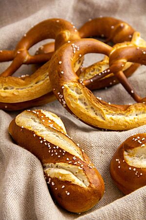 Mehrere Laugenbrezeln und längliche Laugenbrötchen mit brauner Kruste und weißem Salz bestreut liegen auf einem beigen Leinentuch.