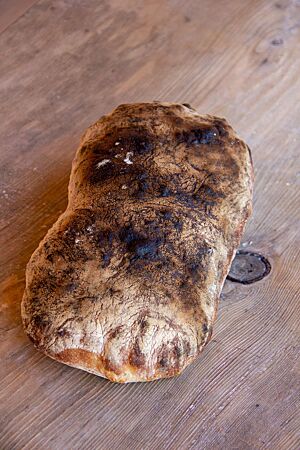 Ein sehr dunkel ausgebackener Laib Ciabatta mit glatter, bemehlter Kruste liegt auf einem Holztisch.