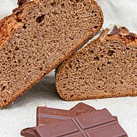 Mehrere Stücke Schokolade liegen vor dem angeschnittenen Schokoladenbrot mit lockerer und elastischer Krume.
