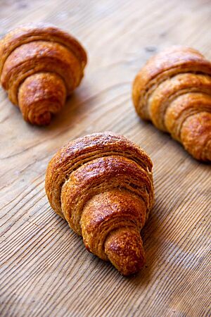 Drei Vollkorncroissants mit glänzender Oberfläche liegen auf einem rustikalen Holztisch.