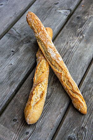 Zwei dünne, lange Stangenbrote mit hellbrauner Kruste und rustikalem Ausbund liegen schräg übereinander auf einem grauen Holztisch.