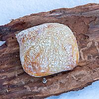Ein goldgelb ausgebackenes, rechteckiges Schneebrötchen mit Mehl auf der Kruste liegt auf einem Stück Baumrinde im Schnee.