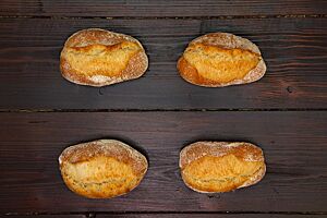 Rustikale Weizenbrötchen. Vier kräftig ausgebackene, oval geformte Weizenbrötchen liegen auf einem Holztisch.