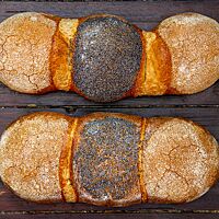 Zwei Weizen-Dinkel-Brote zeigen ihre dreiteiligen Brotlaibe, die mit Grieß und Mohn bestreut sind.