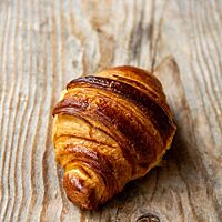 Ein goldbraun glänzendes Croissant mit blättrigen Schichten liegt auf einem Holzbrett.