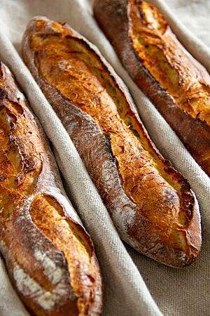 Drei kräftig ausgebackene, rustikal aufgerissene Kartoffelbaguettes mit leicht bemehlter Kruste liegen in einem Leinentuch.