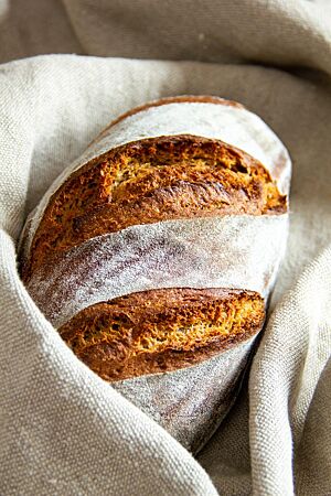 Ruchbrot. Ein goldbraun ausgebackenes Ruchbrot mit bemehlter Kruste liegt in einem Bäckerleinen.