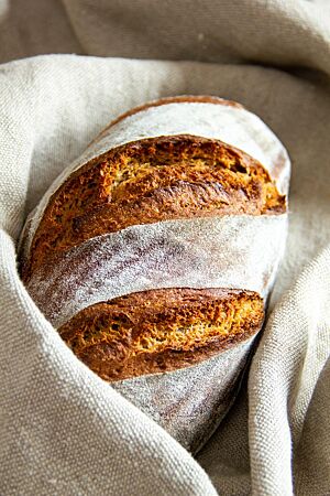 Ein goldbraun ausgebackenes Ruchbrot mit bemehlter Kruste liegt in einem Bäckerleinen.