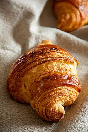 Croissants (2013). Ein goldbraun glänzendes Croissant mit sichtbaren Schichten liegt auf einem Leinentuch.