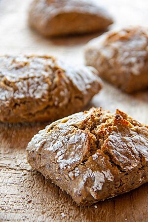 Treberbrötchen mit Gerste. Die rustikal aufgerissenen Treberbrötchen mit leicht bemehlter Kruste liegen auf einem Holztisch.