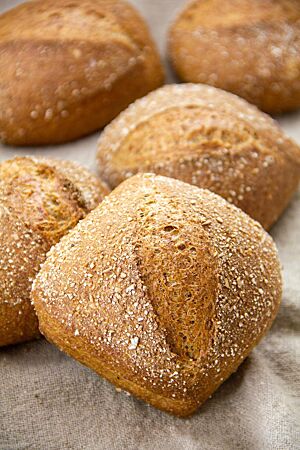 Dinkelschrotbrötchen. Mehrere dunkle, quadratische Brötchen mit diagonalem Einschnitt und Schrot auf der Kruste liegen auf einem Leinentuch.