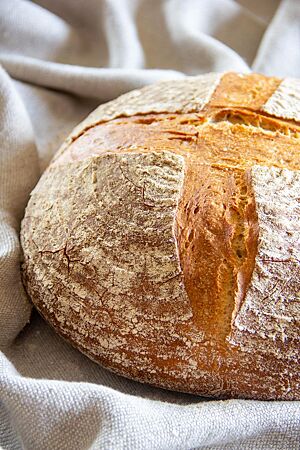 Das goldbraun ausgebackene, kreuzförmig eingeschnittene Bauernbrot mit leicht bemehlter Kruste liegt auf einem Leinentuch.