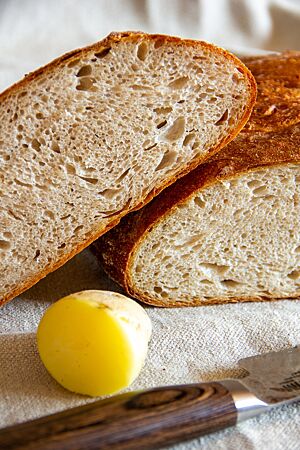 Dinkelkartoffelbrot: luftig und herzhaft. Ein aufgeschnittener Laib Brot mit dunkler, glänzender Kruste und heller, luftiger Krume liegt zusammen mit einer gekochten Kartoffel und einem Brotmesser auf einem Leinentuch.