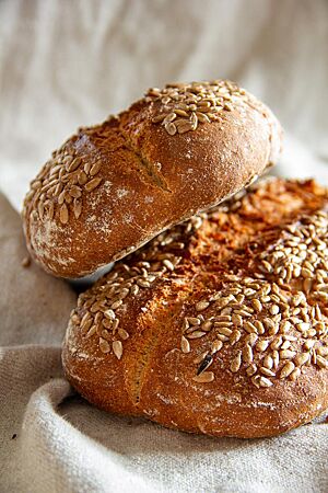 Zwei goldbraun ausgebackene Brotlaibe mit Sonnenblumenkernen auf der Kruste liegen übereinander auf einem Leinentuch.