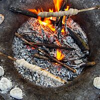 Auf dem Rand einer Feuerschale liegen zehn kleine, stark bemehlte Brötchenteiglinge, während über der mittigen Glut und einigen Flammen Stockbrot gebacken wird.