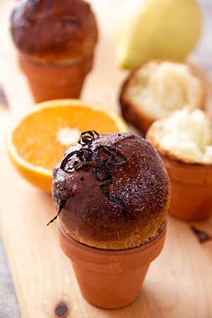 Dinkelbrioche mit Orangen und Zitronen. Auf einem hellen Holztisch stehen mehrere kleine Tontöpfe, die als Backformen für die dunkel ausgebackenen Dinkelbrioches verwendet wurden.