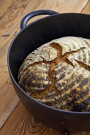 Das kräftig ausgebackene, rustikal aufgerissene Weizensauerteigbrot mit bemehlter Kruste liegt in einem Gusseisentopf.