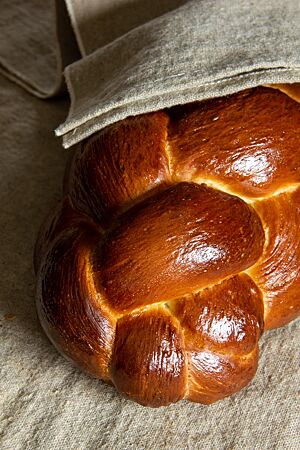Challah (neu). Ein goldbraun ausgebackener Hefezopf mit glänzender Oberfläche liegt in einem Leinentuch.