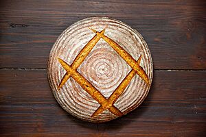 Backesbrot. Ein kräftig ausgebackenes Roggenmischbrot mit leicht bemehlter, eingeschnittener Kruste liegt auf einem Holztisch.