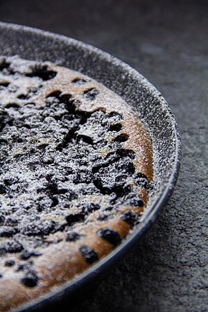 Der fertig gebackene, mit Puderzucker bestreute Getzen liegt in einer runden, flachen Kuchenform. Auf seiner Oberfläche sind Schwarzbeeren (Heidelbeeren) zu sehen.