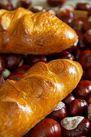 Länglich geformte Eierwecken liegen hellbraun ausgebacken und stark glänzend in einer Holzkiste mit Kastanien.