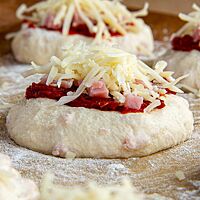 Im rohen Teigling werden Schinkenstückchen sichtbar, ebenso ist auf dessen Oberfläche Tomatenmark, Kochschinken und geriebener Käse zu sehen.