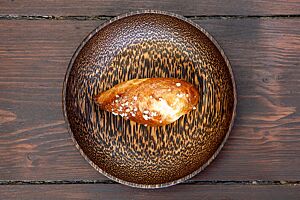 Ein goldbraun ausgebackener, an den Enden spitz zulaufender Brioche-Zipfel mit Hagelzucker auf der Kruste liegt in einer Holzschale.