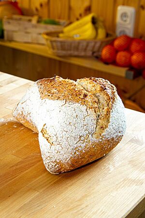 Das goldbraun ausgebackene, rustikal aufgerissene Käppchenbrot mit stark bemehlter Kruste liegt auf einem Holztisch.