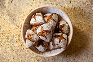 Roggenbrötchen. Die kräftig ausgebackenen, leicht bemehlten Roggenbrötchen mit Kreuzschnitt auf der Oberfläche liegen in einem Gärkorb aus Peddigrohr.