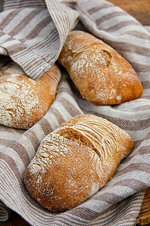 Hefewasser-Dinkelhinkel. Drei leicht bemehlte Dinkelmischbrötchen mit minimal aufgerissener Kruste liegen in einem Leinentuch.
