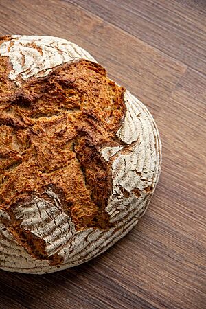 Alles-muss-weg-Brot. Das kräftig ausgebackene, rustikal aufgerissene Alles-muss-weg-Brot mit leicht bemehlter Kruste liegt auf einem Holztisch.