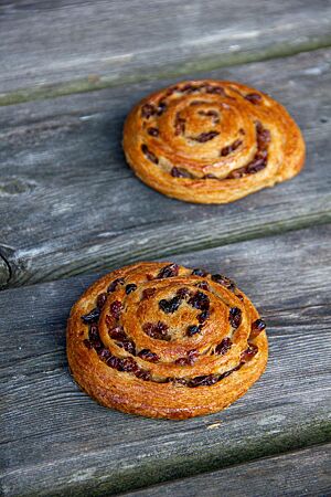Pain aux raisins (Vollkorn und hell). Zwei zur Schnecke geformte, goldbraun ausgebackene Pain aux raisins liegen auf einem rustikalen Holztisch.