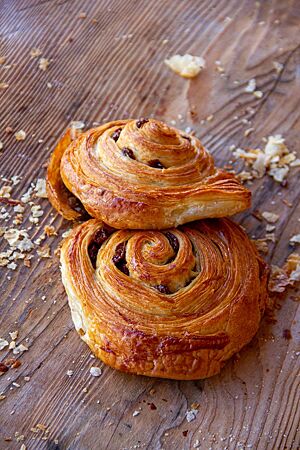 Hüttenplunder (Pain aux raisins). Zwei goldbraun ausgebackene Rosinenschnecken liegen auf einem Holztisch.