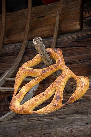 Fougasse. Eine kräftig ausgebackene, schön geformte Fougasse hängt an einem Holzstab vor einer rustikalen Holzwand.
